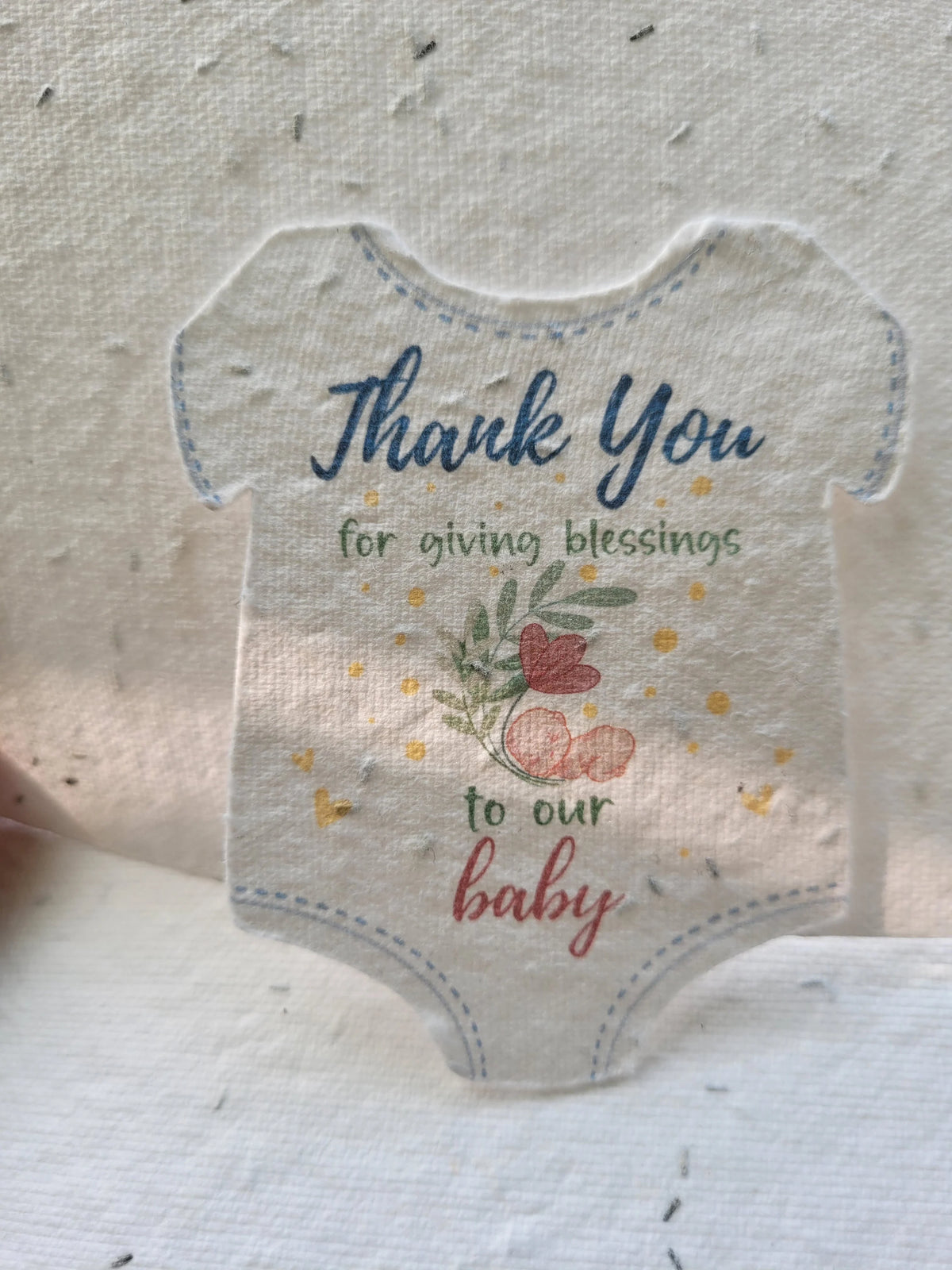 A die-cut fabric ornament shaped like a baby bodysuit, featuring the text 'Thank You for giving blessings to our baby' with a small floral and heart illustration, displayed on a textured fabric background.