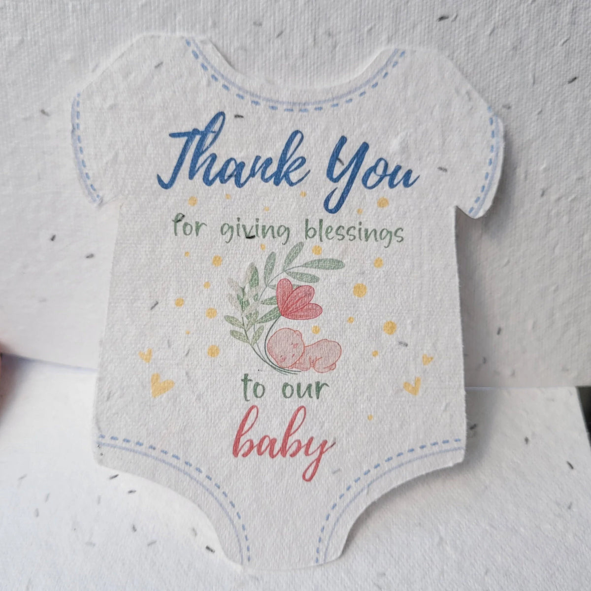 A baby bodysuit-shaped thank you card with a speckled white texture, featuring blue and pink text and a small floral illustration on a textured white background.