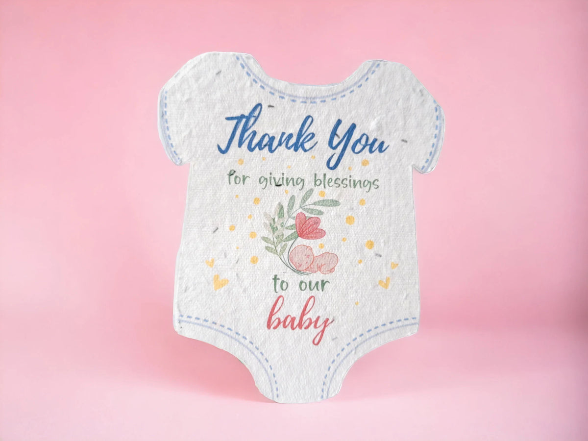 A white short-sleeve baby bodysuit or card-style baby print with blue stitched trim and printed text reading 'Thank You for giving blessings to our baby,' decorated with small hearts, flowers, and a pink baby bottle illustration, shown on a soft pink background.
